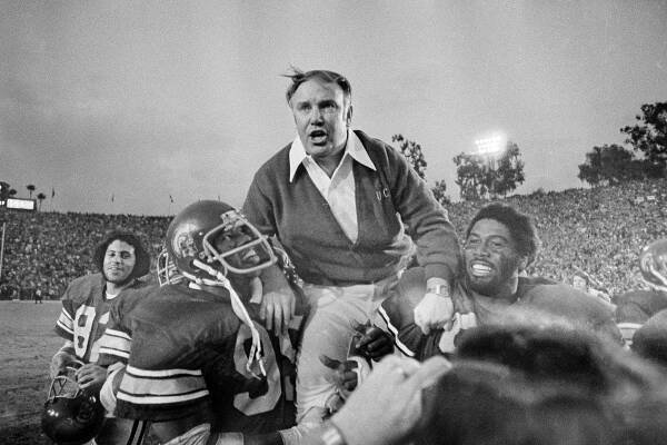A black-and-white photo of John Robinson, a heavyset man wearing a cardigan over a shirt with an open collar, being held aloft by two smiling football players in uniform. Behind them is a view of a packed football stadium.