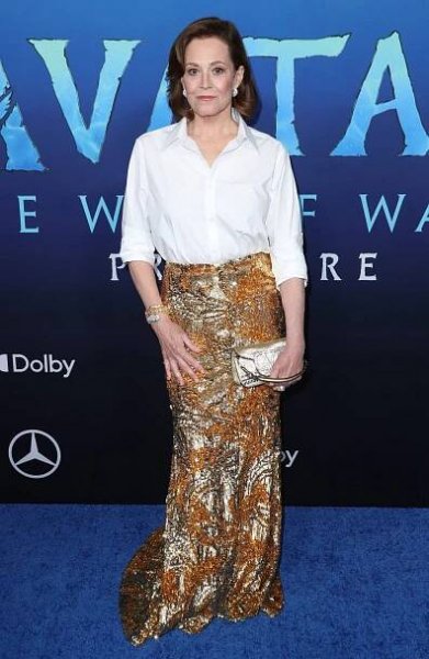 Sigourney Weaver poses in front of a backdrop at an “Avatar” movie event. She wears a button-down white shirt and a long glittery skirt in an abstract bronze and silver pattern. 