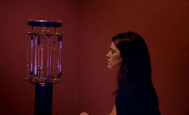 1600 Hernandez, in a burgundy-walled room, stands in profile as she contemplates her father's machine. The machine seems to consist of a cylinder of lit-up glass tubes with a coil on top.