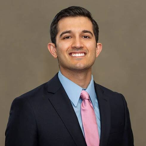 Professional headshot of someone in navy suit and pink tie against neutral background.