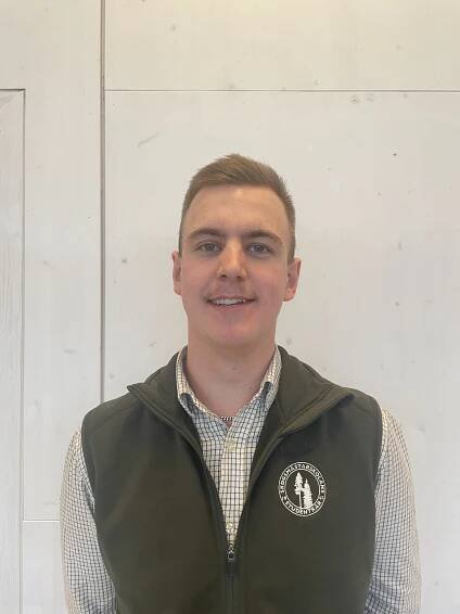 A smiling young man with short dark blonde hair, wearing a blac and white checkered shirt and a black vest with a symbol "Skogsmästarskolans studentkår" in white, stands against a white wall.