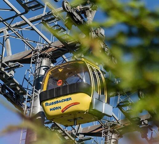 Gelbe Gondel mit dem Schriftzug "Russbacher Horn" hängt unterhalb einer Seilbahnstütze. Rechts im Bild Blätter eines Baumes.