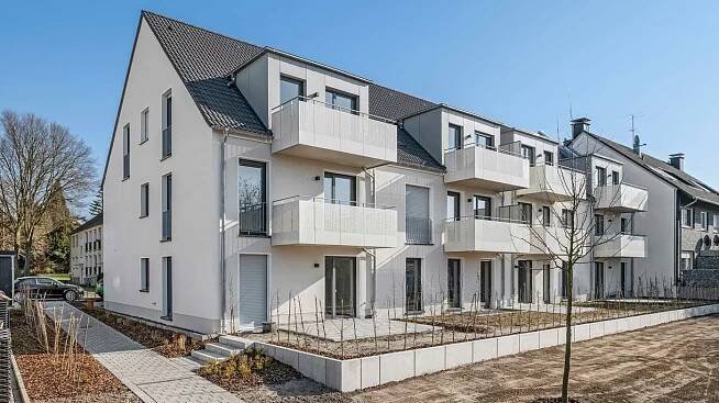 Gartenansicht eines VIVAWEST-Gebäudes in Mülheim an der Ruhr. Das Haus besitzt ein graues Spitzdach mit drei großen Gauben und acht grau verkleideten Balkonen. Im Erdgeschoss sind die neu angelegten Terrassen und Gartenflächen zu sehen. Die Gärten sind von einer neu gesetzten Hecke umgeben. Davor befindet sich eine planierte Freifläche, auf dem später Rasen eingesät wird.  