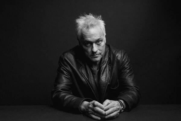 Black-and-white portrait of Marc Ribot, an older white man with white, wispy hair. He is wearing a leather jacket and has his elbows on the table in front of him.