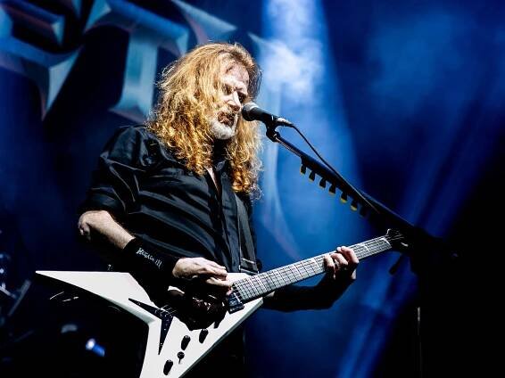 Dave Mustaine on stage, he has a white V-shaped guitar in hand and stands before the mic, with blue lighting in the background.