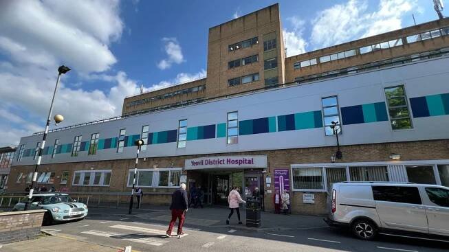 People walking into Yeovil District Hospital. The building has brown bricks and purple lettering saying 'Yeovil District Hospital'. There are cars outside and a zebra crossing.