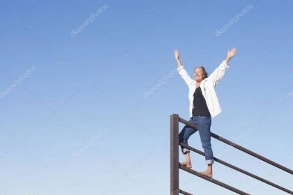 Stáhnout - Portrét atraktivní starší ženy stojící v vítězné póze nad kovovým plotem, šťastný úsměv, paže vzhůru a radostná, s modrou oblohou jako pozadí a kopírovací prostor. — Stock obrázek