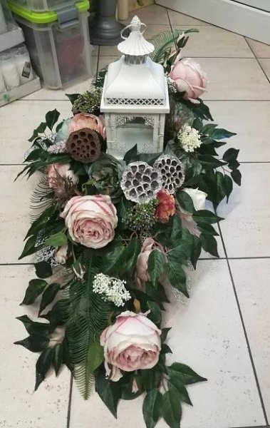 a white lantern surrounded by pink flowers and greenery on the floor in front of a trash can