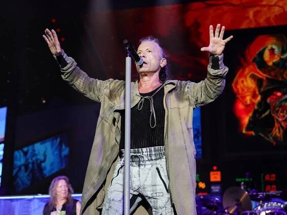Bruce Dickinson on stage. He is singing into a mic on a stand, and has both hands in the air.