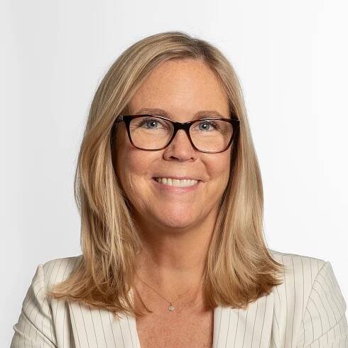 A woman with shoulder-length blonde hair and glasses smiles. She is wearing a white pinstripe blazer.
