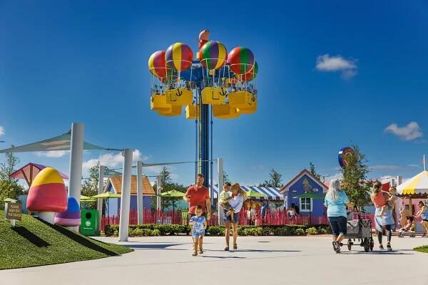 A group of families enjoys the Fun Fair attraction at Peppa Pig Theme Park in Dallas Fort Worth, playing games and riding various attractions. In the background, Peppa's Balloon ride can be seen, adding to the vibrant and playful atmosphere of the park.