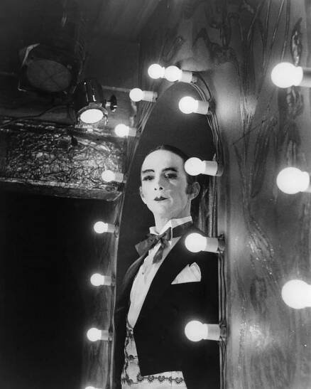 A man with slicked-back black hair, his face covered in white makeup, looks sternly into a round mirror that’s ringed with lightbulbs.