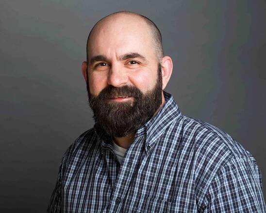A bald man with a beard is wearing a plaid shirt and smiling.