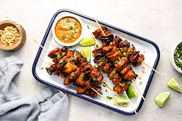 white serving platter with cooked chicken skewers and small bowl of peanut sauce