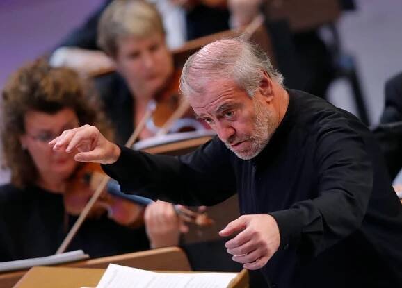 Valery Gergiev, wearing a black jacket, conducts an orchestra. Violinists can he seen in the background, out of focus.