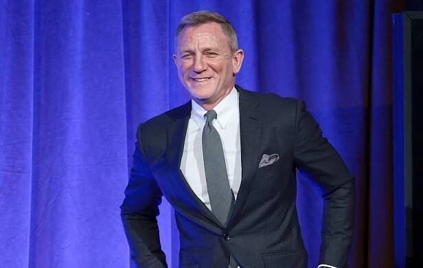 Daniel Craig in a tailored suit with a pocket square, smiling against a curtain backdrop
