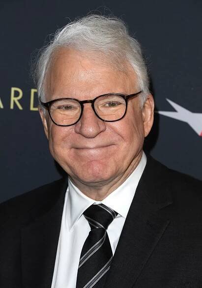 Steve Martin wearing a black suit and tie, smiles at the camera at a celebrity event