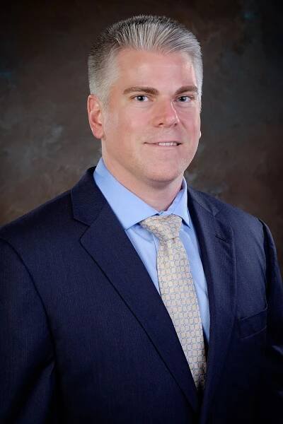 Professional headshot of a man with a clean-shaven look, wearing a navy suit and blue shirt, against a minimalist studio setting.