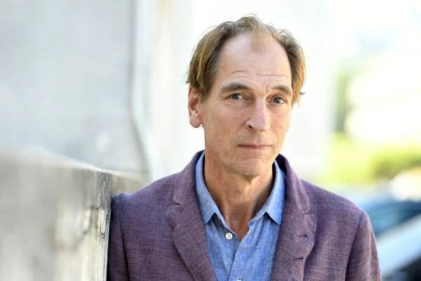 Julian Sands, wearing a blue dress shirt and purple blazer, stares at the camera.