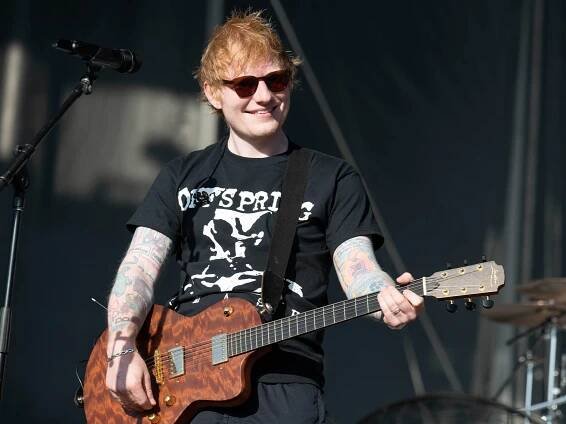 Ed Sheeran on stage in 2024. He is wearing sunglasses and is smiling. He has an electric guitar in hand.
