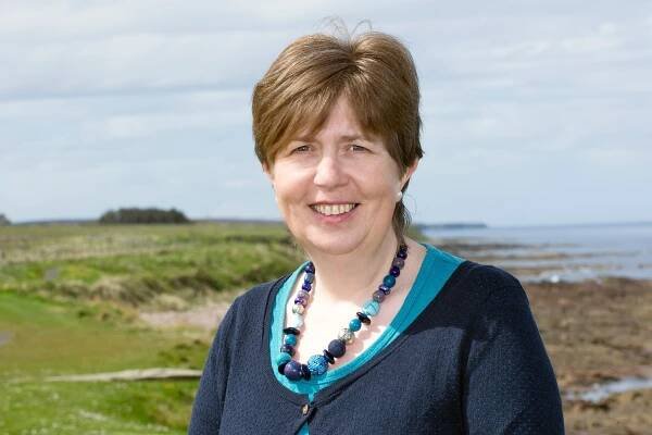 Kate Buchanan smiles at the camera in a rural landscape with sea in the background. She has short brown hair and wears a black shirt with chunky necklace.