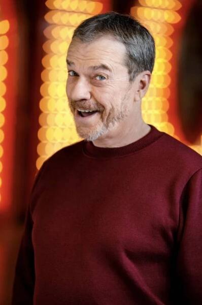 Man smiling with a beard, wearing a plain crewneck sweater, standing in front of a blurred background with circular light patterns