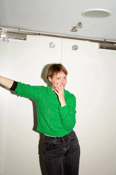 Katharina Marie Schubert is standing in front of a white wall in a green sweatshirt. She smiles behind her left hand.