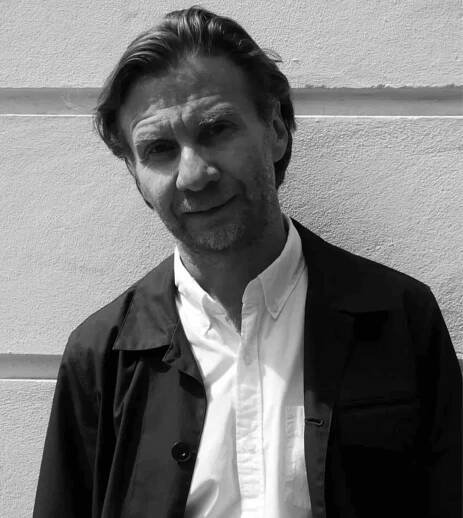 A man with medium-length hair and a beard wearing a dark jacket over a white shirt stands against a textured wall. Black and white photo.