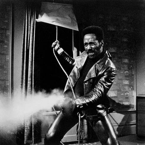 Richard Roundtree, who is Black, fires a gun while pulling a rope. He is wearing a leather jacket and pants and has a mustache.