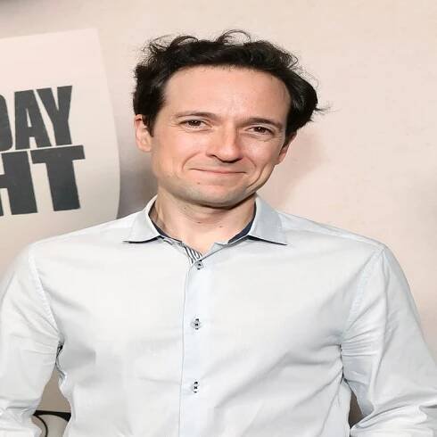 A man in a white shirt and jeans stands in front of a backdrop featuring "Saturday Night" posters