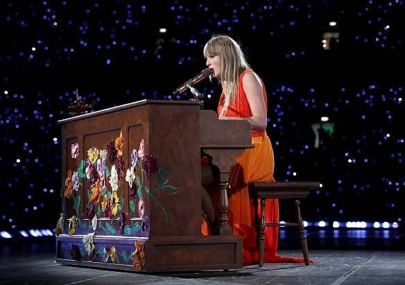 A person plays a floral-decorated piano on stage in a stadium with a glowing crowd in the background