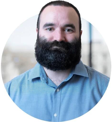 David Naughten smiling at the camera. Heh as cropped black hair with a large black beard and brown eyes.