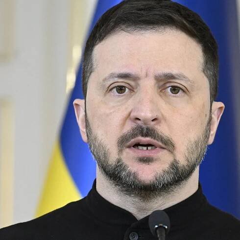 This photo shows Ukrainian President Volodymyr Zelenskyy photographed from about the shoulders up. He's wearing a black shirt and has a dark beard. Behind him is a blue and yellow Ukrainian flag.