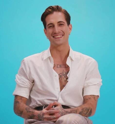 Damiano David, with tattoos and slicked-back hair, smiles, seated, wearing a casual button-up shirt