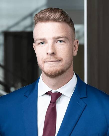 A man with short, light-colored hair and a beard is wearing a blue suit, white shirt, and a maroon tie. He is standing indoors with a blurred background.