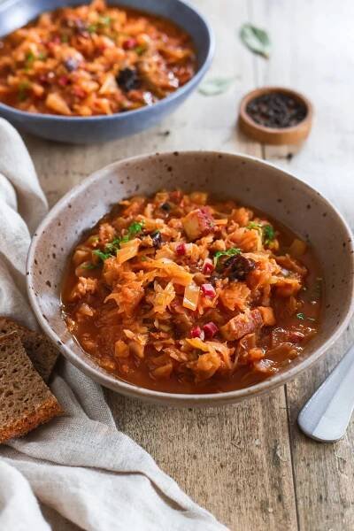 Veganes Bigos in einer rustikalen Keramikschüssel, serviert mit frischem Brot auf einem Holztisch. Polnischer Krauteintopf mit Sauerkraut, Pilzen und Gewürzen, perfekt für eine herzhafte Mahlzeit.