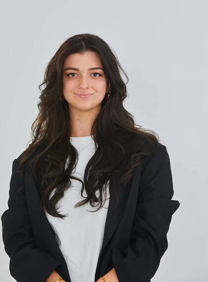 Young woman with long dark hair in a black blazer and white shirt, smiling against a light gray background.