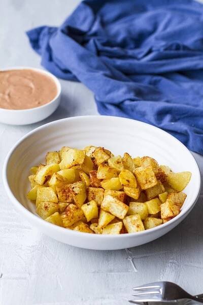 Large white bowl with fried cubed potatoes covered in chili spices. Blue linen in the background.