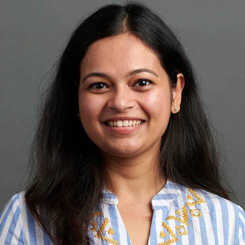 A person with long dark hair smiles at the camera, wearing a white and blue striped shirt with yellow embroidery, against a gray background.