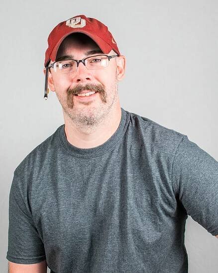 Smiling person with a mustache and red baseball cap poses with a pencil behind his ear