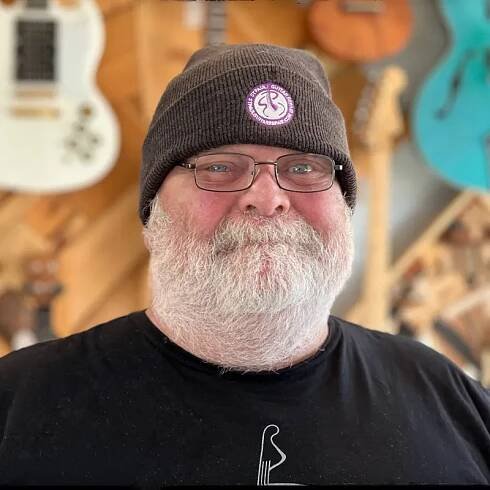 Portrait of John Tracy, smiling in front of a wall of custom built guitars