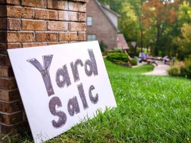 Yard sale sign sitting on grass in front of brick home.