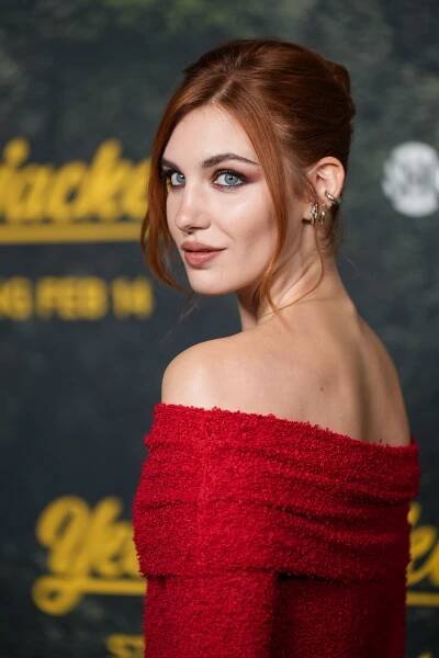 Person with styled auburn hair in an elegant off-the-shoulder red dress, posing at a film event backdrop
