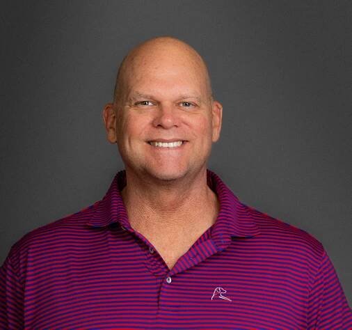 A bald man wearing a purple and blue striped polo shirt is smiling for the camera.