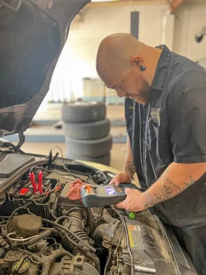 A mechanic with tattoos on his arms and a black uniform with a name tag works under the hood of a car in a garage. 