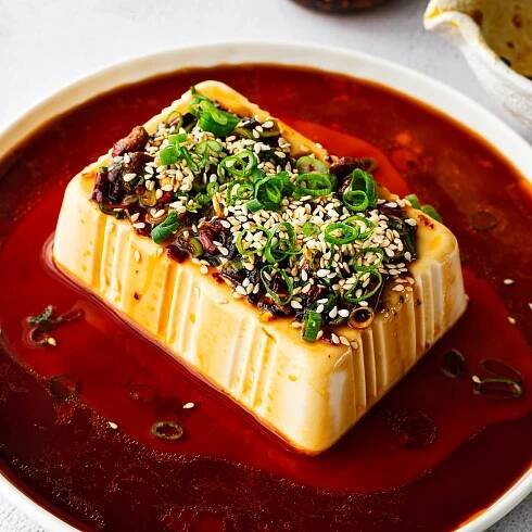 A block of silken tofu surrounded by a soy-based dressing with a soy sauce bowl and a bottle of chili crisp visible in the back ground.