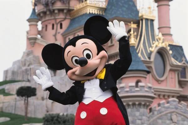 Mickey Mouse in a tuxedo waving in front of Sleeping Beauty Castle at Disneyland Paris.