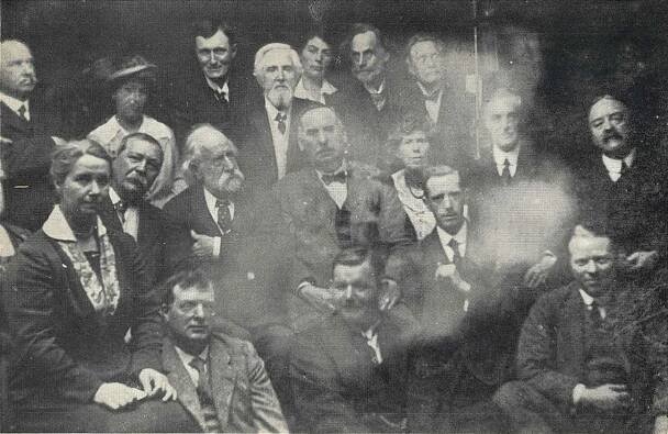 photogrpah of a group of men and women seated. in the centre of the image on its side is the ghostly face of a man