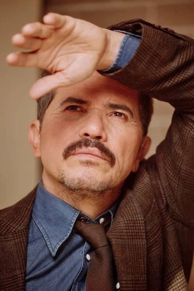 A portrait of a man who is wearing a brown blazer over a blue jean collared shirt and brown tie. He is resting his head against his raised left arm.