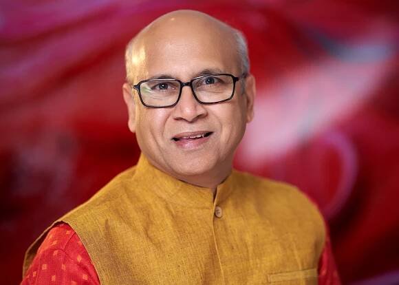 Portrait of someone wearing glasses and a golden yellow kurta against a red backdrop.
